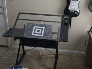 Newly assembled black adjustable art desk with a tilted glass work surface, gridded cutting mat, side organizers, and an electric guitar hanging in the background.