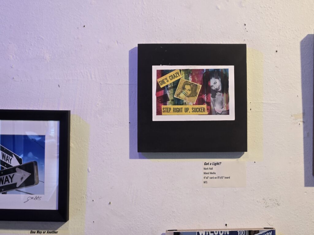 Exhibition display of the mixed-media collage Got a Light? by Mark Hall hanging on a white textured gallery wall. The horizontal 4x6 artwork is matted on a square black mount. The collage layers colorful, textured street-art and print elements behind vintage pop-culture imagery, including a woman drinking from a cup and a portrait of a smiling woman. Bold yellow headline strips accent the piece, reading "SHE'S CRAZY" at the top left and "STEP RIGHT UP, SUCKER" across the bottom. A printed gallery label is affixed to the wall below the artwork.