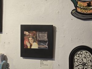 Mark Hall's mixed-media collage Just a Bad Dream mounted on a black square board hanging on a gallery wall alongside other artwork, featuring a vintage portrait of a woman, a retro "SEXORAMA" newsprint clipping, and an exhibition label below.